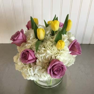 Compact arrangement of yellow tulips, lavender roses, and white hydrangeas in a clear glass vase
