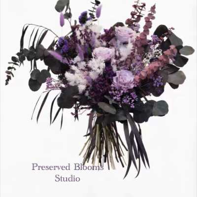 Hand-tied bouquet of preserved purple, lavender, and white flowers with dark foliage.