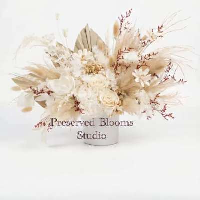 Low arrangement of preserved neutral-toned flowers with roses and dried grasses in a white cylinder vase.
