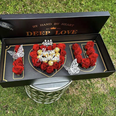 Red roses arranged in a black gift box with gold accents and a "Happy Anniversary" topper