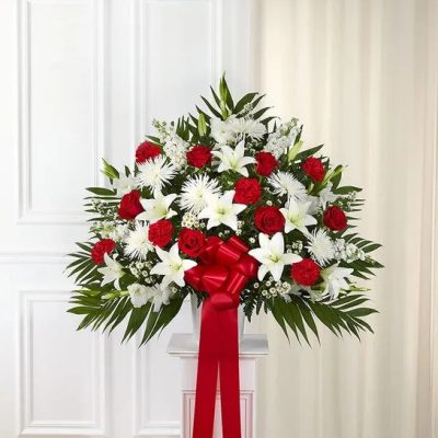 Fan-shaped red and white arrangement with roses, lilies, and mums on a pedestal with a red bow