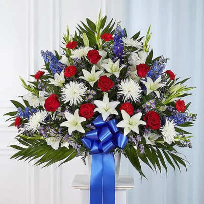Large red, white, and blue standing floral arrangement with lilies, roses, and a blue ribbon bow