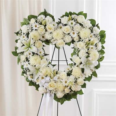 Heart-shaped standing wreath of white flowers with a ribbon on a metal easel.