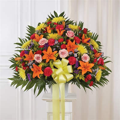 Large fan-shaped arrangement of orange lilies, pink roses and mixed bright flowers in a white urn with yellow bow