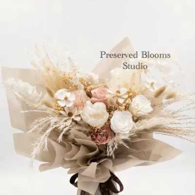 Bouquet of preserved white and blush flowers with dried grasses wrapped in tan paper and tied with a brown ribbon