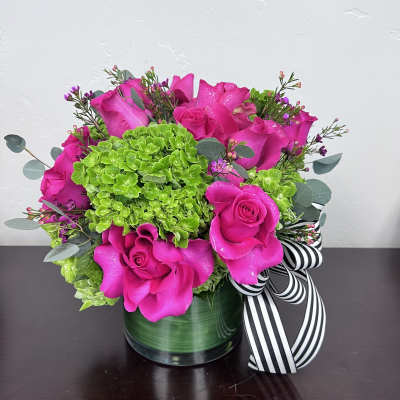 Compact arrangement of hot pink roses and green hydrangeas in a short glass vase with a striped ribbon bow.