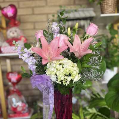Pink lilies and roses arranged in a tall magenta vase with a purple ribbon