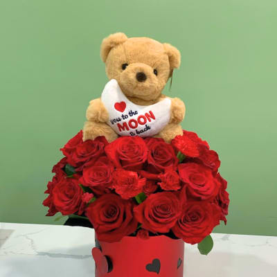 Teddy bear holding a heart pillow above red roses in a heart-decorated box