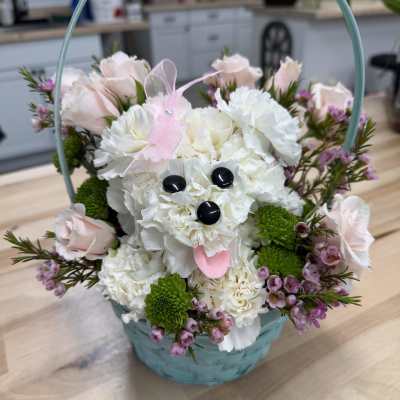 Basket arrangement with a white carnation dog surrounded by pink roses and small purple flowers.
