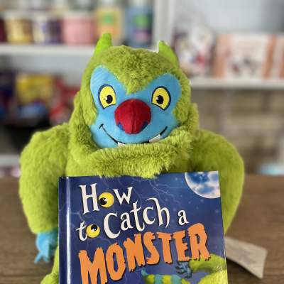 A green plush monster holding a children's book titled "How to Catch a Monster."
