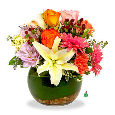 Colorful arrangement of lilies, roses, gerbera daisies, and carnations in a round green glass vase with pebbles.