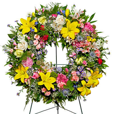 Colorful standing wreath of yellow lilies, pink carnations, and mixed blooms on a metal easel.
