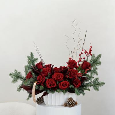 Red roses arranged in a white vase with pine branches and berries