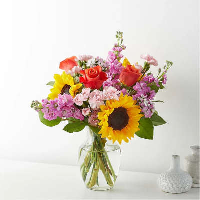 Mixed bouquet of sunflowers, roses, and pink blooms in a clear glass vase