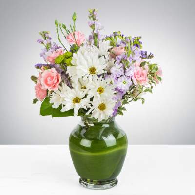 Pastel bouquet of white daisies, pink carnations, and lavender flowers in a clear vase lined with green leaves