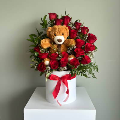 Round box of red roses with small white blooms and a teddy bear nestled in the center.