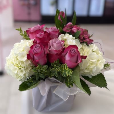 Compact arrangement of pink and lavender roses with white hydrangeas in a square vase with ribbon.
