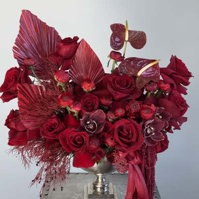 Deep red rose arrangement in a silver urn with burgundy tropical accents