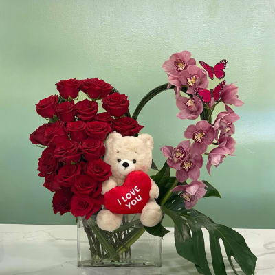 Red roses and pink orchids in a glass vase with a teddy bear holding a heart