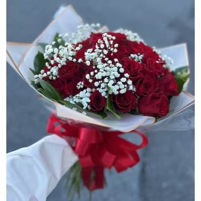 Handheld bouquet of red roses with white filler flowers wrapped in paper and tied with a large red bow.