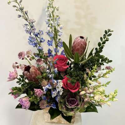 Tall mixed bouquet with pink roses, blue delphinium, and protea in a square vase