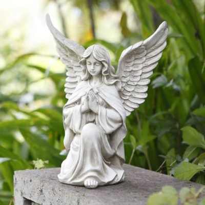 White kneeling angel statue with wings on a stone ledge in a lush garden.