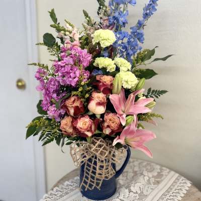 Mixed bouquet of pink, blue, and yellow flowers in a blue pitcher vase