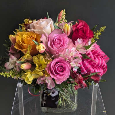 Compact arrangement of pink, yellow, and red roses with alstroemeria in a square glass vase with pink ribbon.