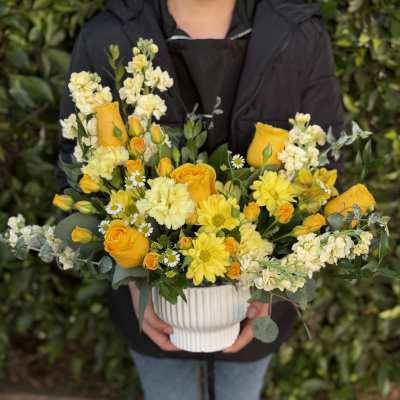 Yellow and cream flower arrangement with roses and daisies in a white ribbed ceramic pot