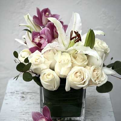 White roses and lilies arranged in a square glass vase with purple orchids