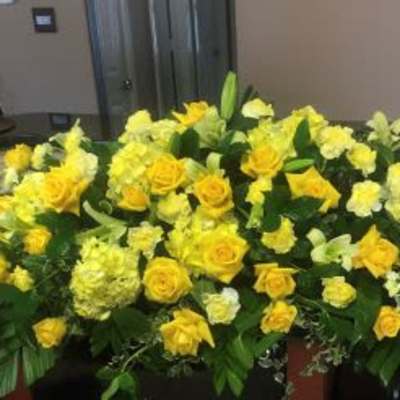 Elongated spray of yellow roses and pale yellow flowers with lush foliage in a low, casket-style arrangement