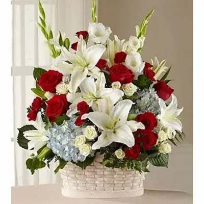 Tall basket arrangement of white lilies, red roses, and blue hydrangeas in a white woven basket
