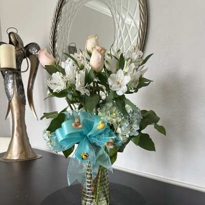 Vase arrangement of white alstroemeria, pale pink roses, and blue hydrangea with a teal bow and small baby-themed decorations.