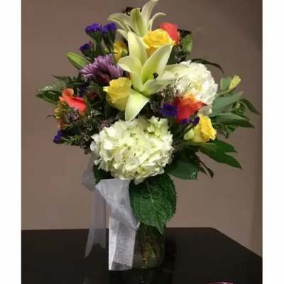 Mixed bouquet of white hydrangeas, lilies, yellow and orange roses in a clear glass vase with ribbon