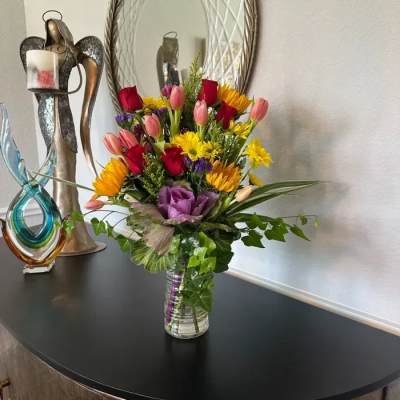 Tall mixed bouquet with red roses, pink tulips, yellow daisies and purple accents in a clear glass vase