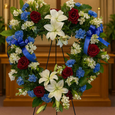 Heart-shaped floral wreath with red roses, white lilies, and blue accents on an easel