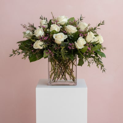 Arrangement of white roses with pink filler flowers in a clear glass cube vase against a pink background