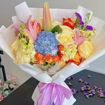 Bouquet of roses, lilies, and blue hydrangea wrapped in white paper with a purple ribbon