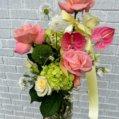 Tall arrangement of pink roses, green hydrangea, and pink anthurium in a clear vase with yellow ribbon