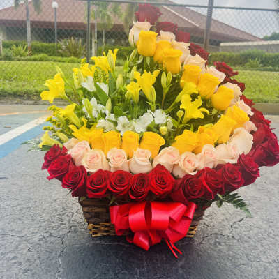 Basket arrangement of red, yellow, and blush roses with lilies and a red bow