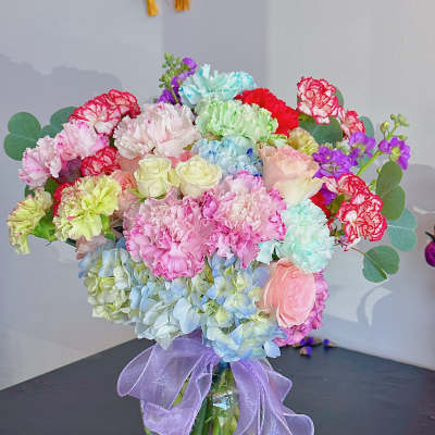 Colorful bouquet of carnations, roses, and hydrangeas in a glass vase with a purple ribbon