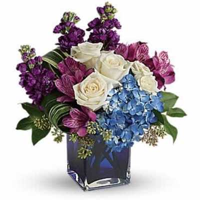 Compact bouquet of white roses, blue hydrangea, and purple flowers in a blue glass cube vase