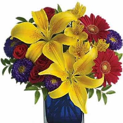 Bright mixed arrangement with yellow lilies, red gerberas, and purple blooms in a blue glass cube vase