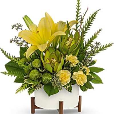 Low arrangement of yellow lilies, carnations, and green blooms in a white pot on a wooden stand
