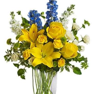 Tall mixed arrangement of yellow lilies and roses with blue and white flowers in a clear glass vase