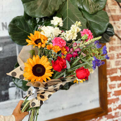 Handheld bouquet of sunflowers, roses, and mixed blooms wrapped in polka-dot paper