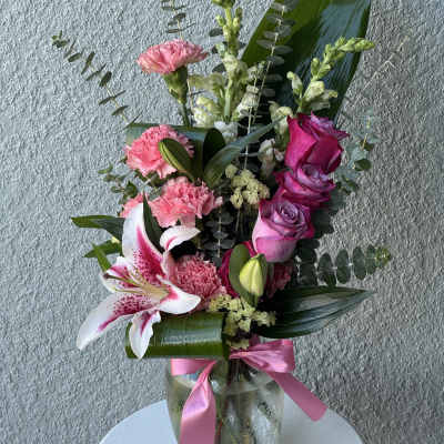 Tall pink and white arrangement with roses, lilies, and carnations in a glass vase with a pink ribbon.