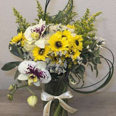 Yellow sunflowers and white orchids in a glass vase with a birthday ribbon