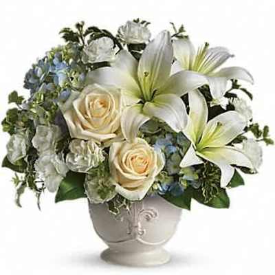 Arrangement of white lilies, ivory roses, and blue hydrangeas in a white urn vase