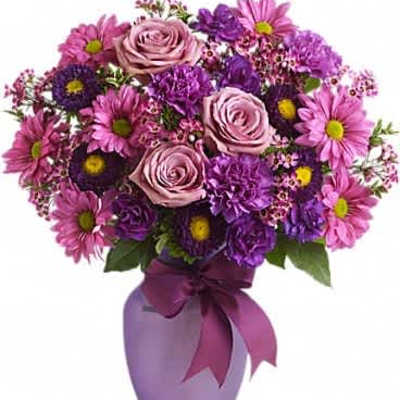 Purple and pink mixed bouquet of roses, carnations, and daisies in a frosted lavender vase with bow